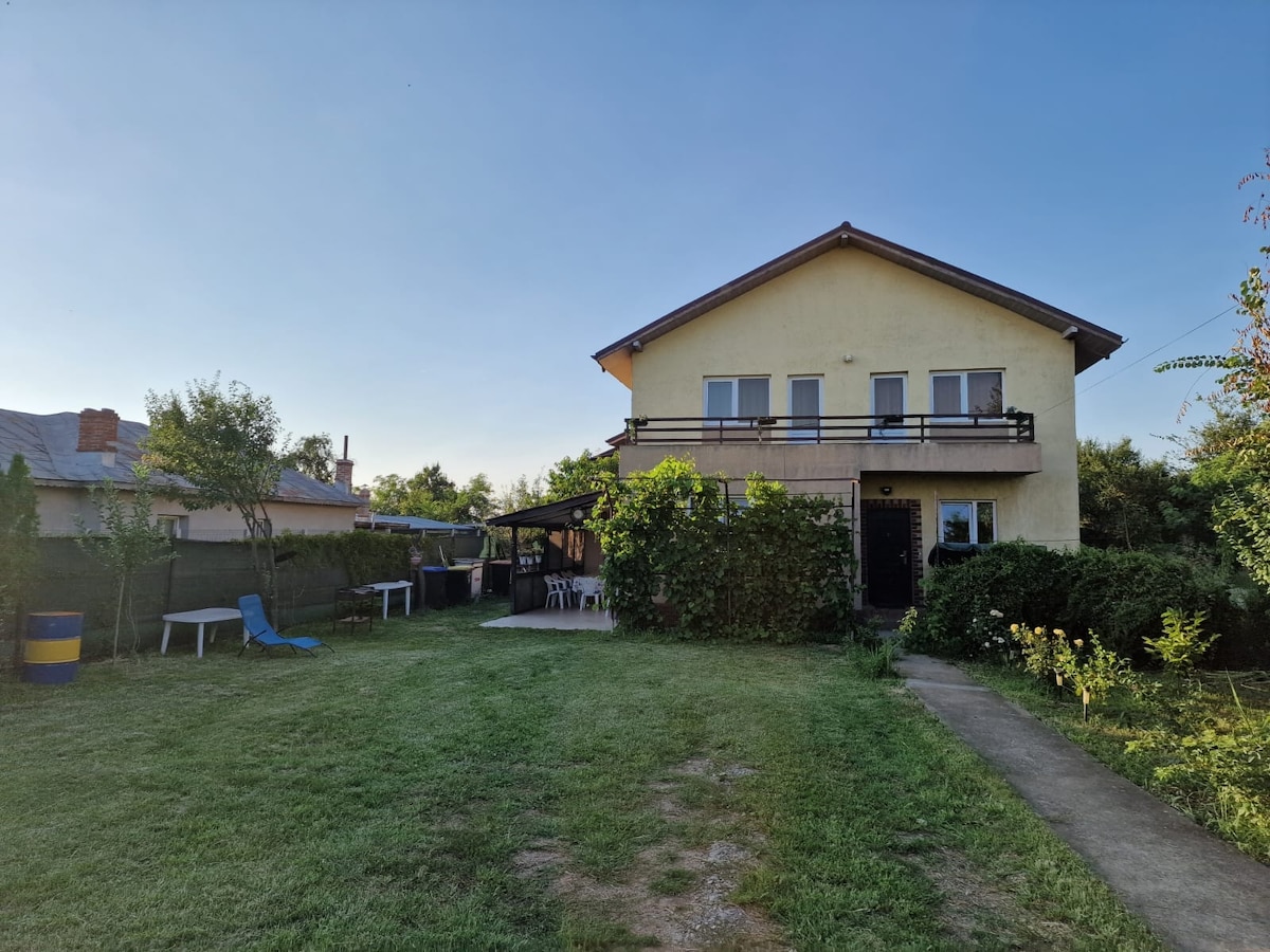 A two-story villa is set in a spacious yard, featuring a well-maintained lawn. Outdoor seating and a shaded area are visible, surrounded by various greenery. The house boasts a balcony and multiple windows, allowing for plenty of natural light.