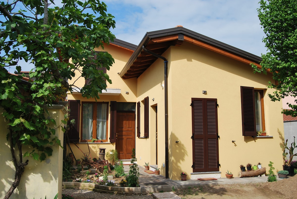 The exterior of the building showcases a yellow facade with wooden shutters. Two windows, adorned with plants, flank the entrance. The welcoming doorway is framed by an inviting garden area featuring various plants. The roof slopes gently, reflecting a traditional design.