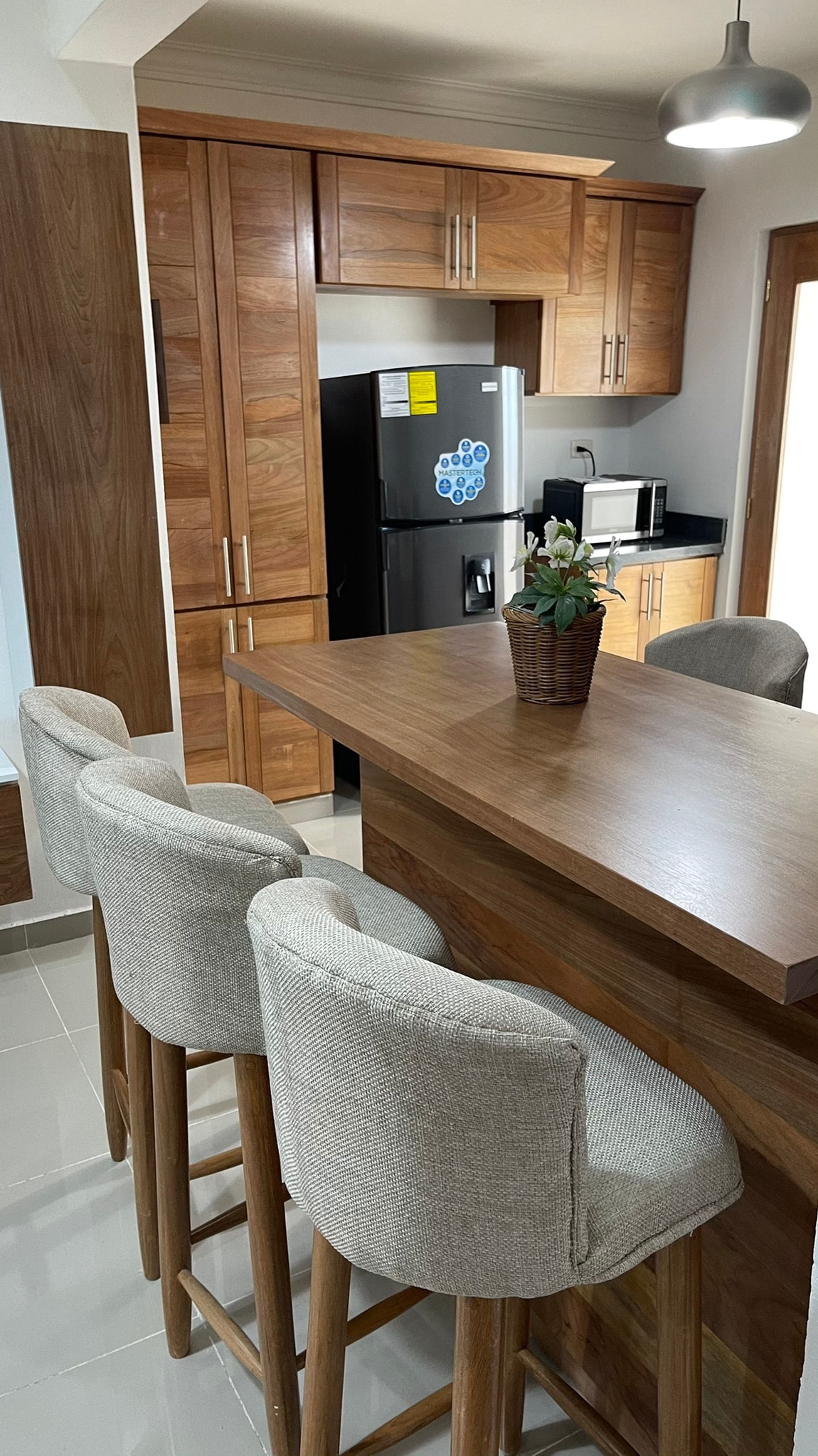 A modern kitchen is presented with warm wooden cabinetry and a contrasting dark countertop. Four bar stools with soft, light-colored upholstery are positioned at a large island. A potted plant adds a touch of greenery, complementing the sleek appliances and minimalistic design.