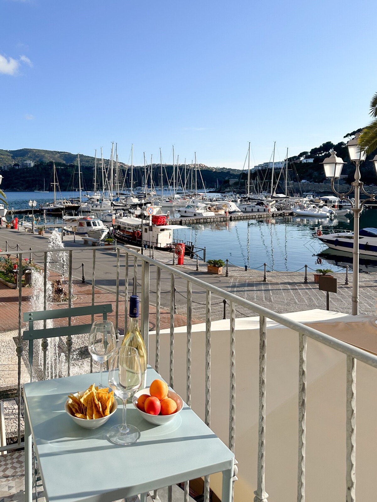 A small table is set for two on a balcony, displaying a plate of snacks and fresh fruit. The view captures a beautiful marina filled with boats, framed by hills under a clear blue sky.