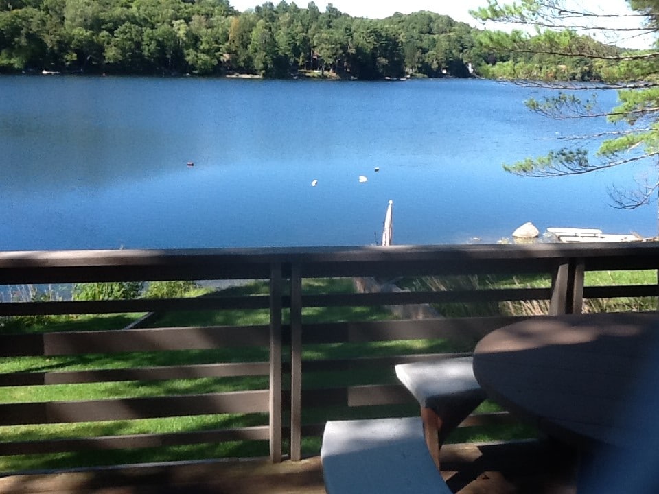 A serene view of Beebe Lake is captured from a deck, showcasing still water reflecting the surrounding greenery. A small table stands on the deck, with wooden railings framing the tranquil scene that includes distant boats dotting the lake's surface.