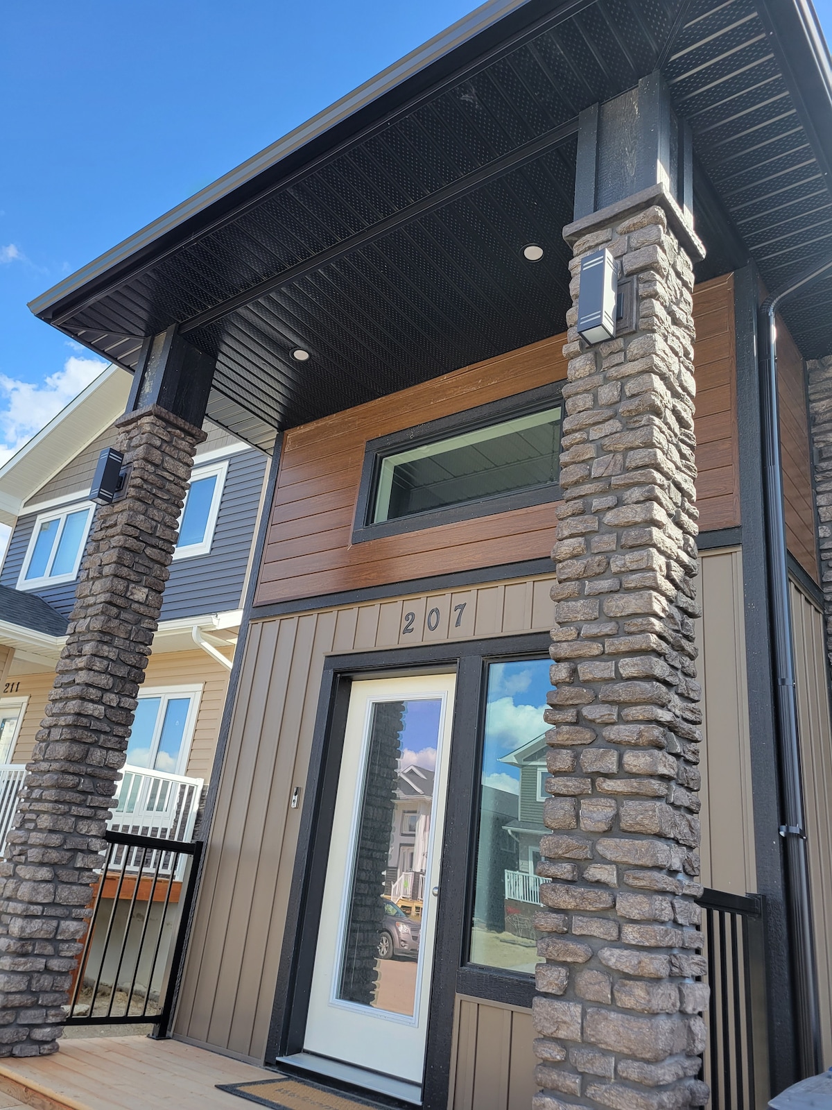 The exterior of the home features a welcoming entrance with a silver door, framed by tall stone columns. The facade includes a mix of wood and vertical siding, creating a modern look. Clear blue skies highlight the architectural details of the structure.