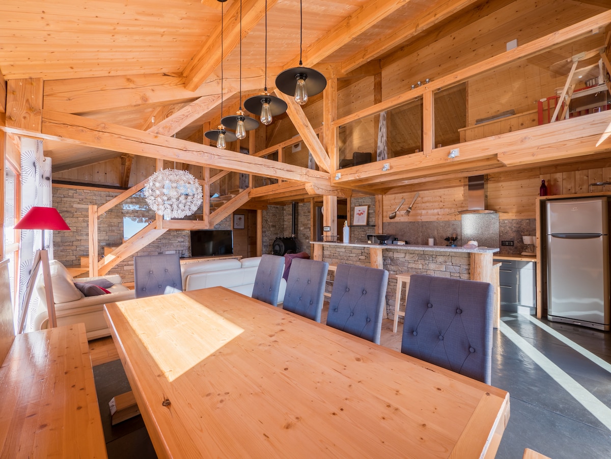 A spacious dining area features a large wooden table surrounded by elegant grey upholstered chairs. A modern light fixture hangs overhead, while wooden beams contribute to the rustic charm. The open layout connects the dining space with a cozy seating area and kitchen, enhancing accessibility.