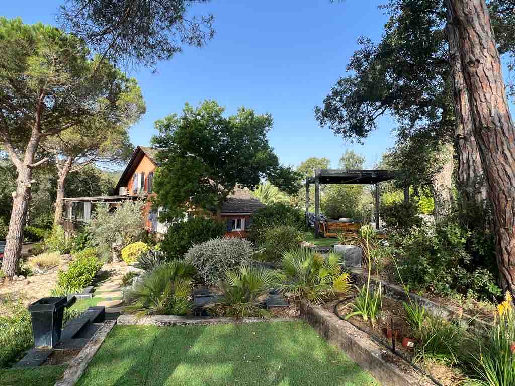 A well-maintained garden is surrounded by tall trees and features a variety of plants. A cozy house is visible in the background, offering shaded seating areas. The garden blends greenery with pathways, creating a calm outdoor space for relaxation and family activities.
