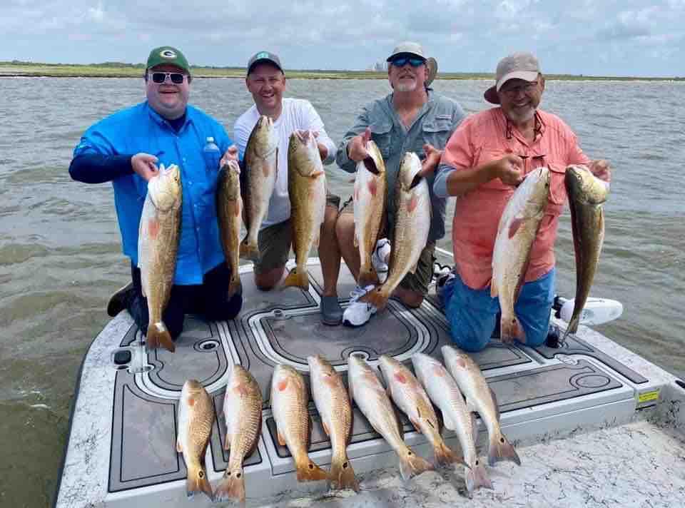 A group of people is seen standing on a boat holding several large fish. The catch is displayed on the deck, showcasing a variety of sizes. The water surrounds the boat, reflecting the cloudy sky above, while the distant shoreline is faintly visible.