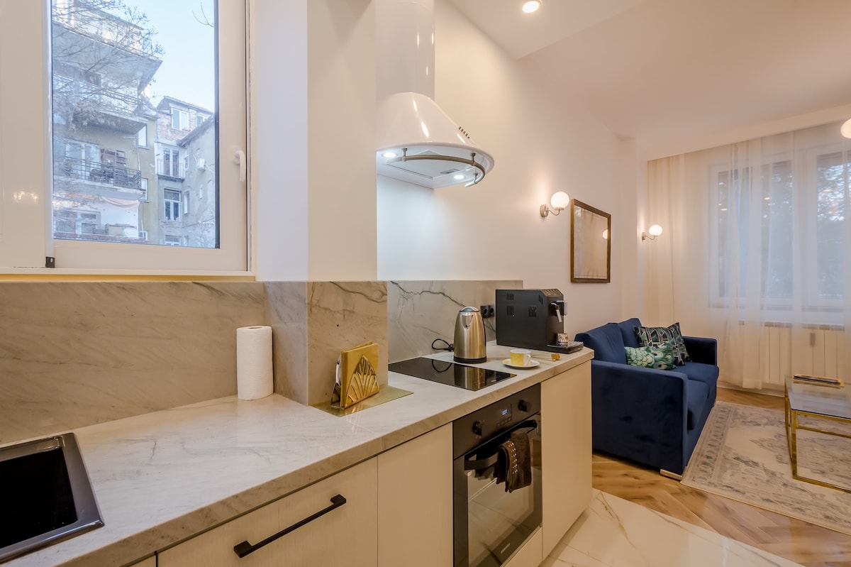 A modern kitchen features sleek granite countertops, luxury appliances, and a cooking area with a built-in stovetop and oven. Natural light filters through the window, illuminating a cozy seating area with a blue sofa and decorative cushions.