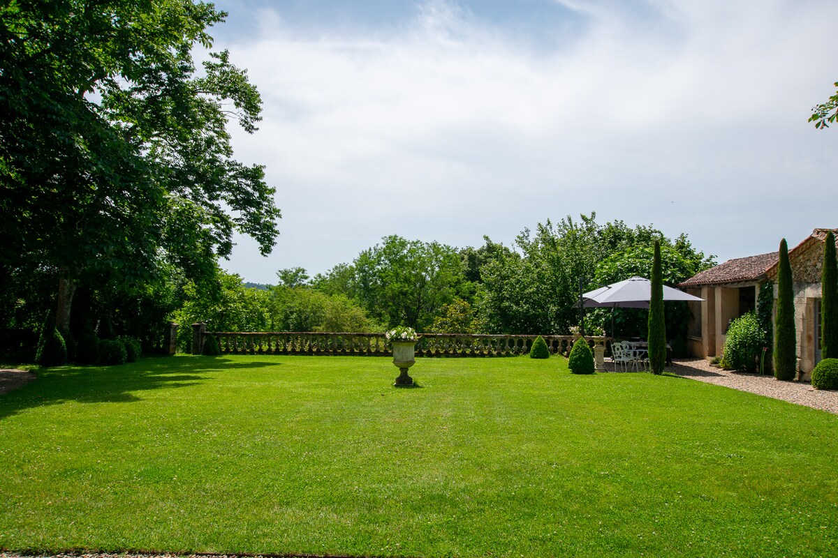A spacious lawn is seen, surrounded by lush greenery and trees. A decorative stone planter stands at the center, while a shaded area with an umbrella provides a comfortable seating spot. The garden's peaceful setting invites relaxation and outdoor gatherings.