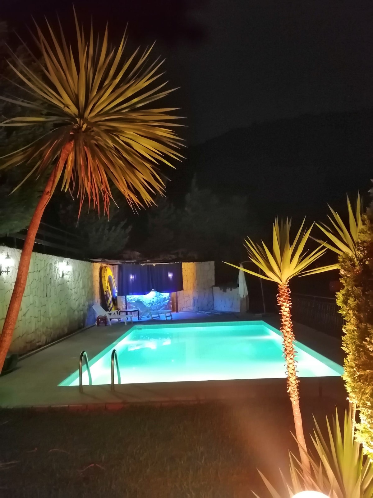 A well-lit swimming pool is surrounded by greenery, with palm trees adding a tropical touch. Soft lights reflect on the water's surface, creating a serene ambiance. Comfortable seating is visible nearby, making the space inviting for relaxation.
