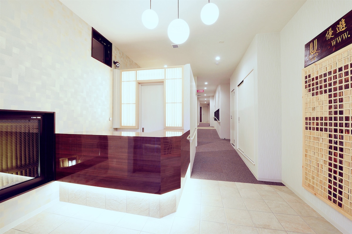 The image depicts a well-lit hallway featuring smooth tile flooring and neutral-toned walls. A reception area with a contemporary wooden counter is visible, alongside illuminated circular light fixtures. The corridor extends beyond, leading to various rooms, creating a welcoming ambiance.