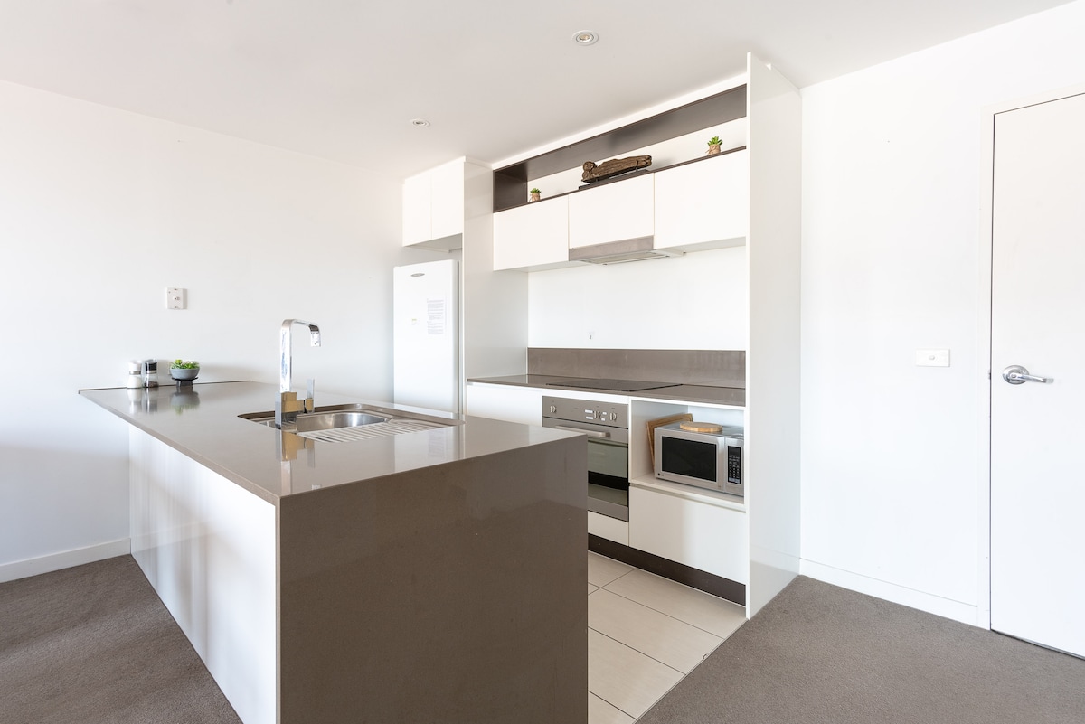 A modern kitchen is equipped with a central island and minimalist cabinetry. Stainless steel appliances, including an oven and microwave, are integrated into the design. Neutral tones create a sleek aesthetic, complemented by a subtle decorative plant on the countertop.