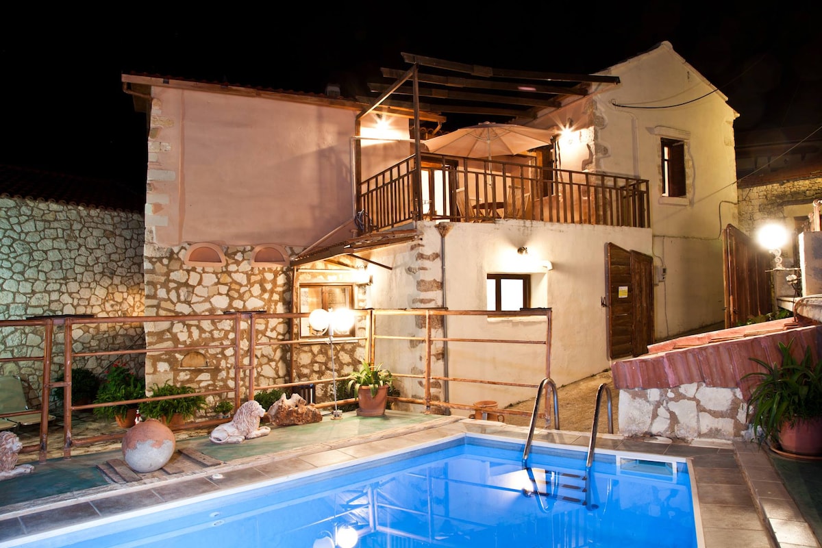 The exterior of the villa is illuminated gently at night, highlighting its combination of traditional and modern architectural features. A private pool is visible in the foreground, with steps leading into the water. The upper balcony offers an inviting space, adorned with an umbrella and chairs.