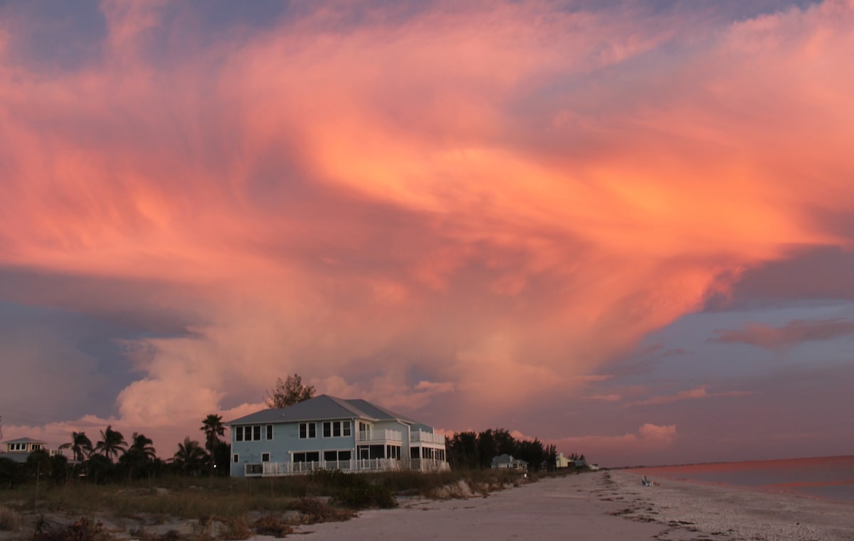 A beachfront townhouse is positioned along a sandy shore, with large windows reflecting the vibrant colors of a sunset sky. Soft clouds in shades of pink and orange create a serene backdrop over calm waters, suggesting a peaceful evening by the Gulf of Mexico.