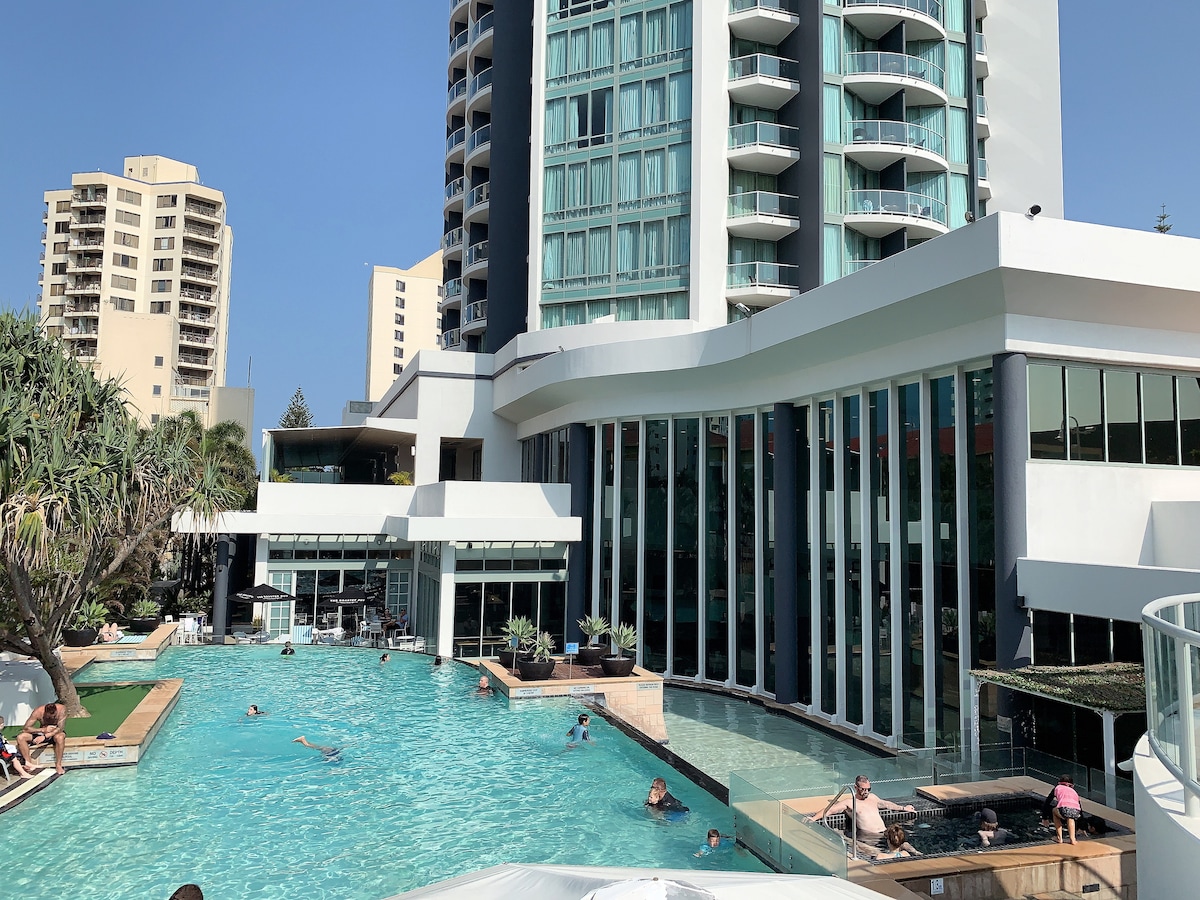 An outdoor swimming pool is featured at the hotel, surrounded by tall palm trees and lounge areas. The building's glass facade reflects sunlight, enhancing the spacious feel. Guests can be seen enjoying the water, with a poolside seating area visible in the background.