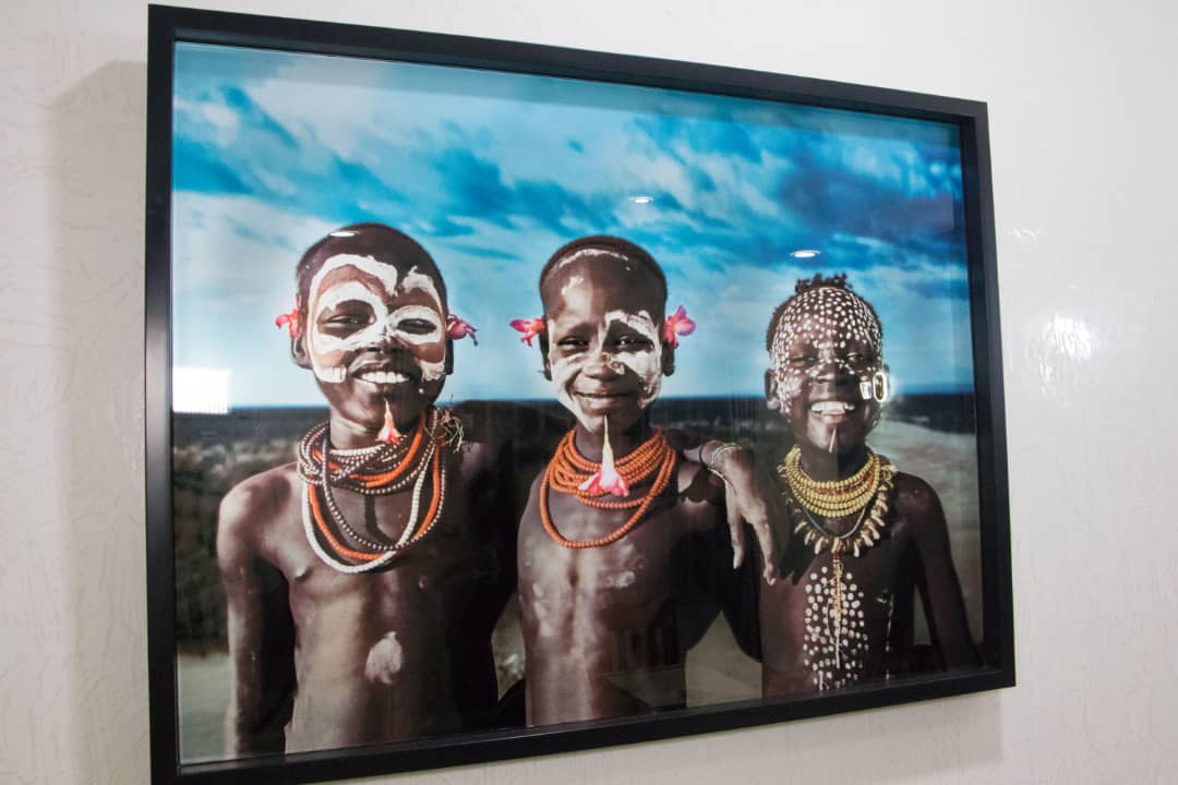 A framed photograph features three individuals adorned with traditional jewelry and colorful accessories. The backdrop displays a sky with clouds, contributing to the cultural essence of the artwork. The subjects' faces are obscured, ensuring privacy while highlighting the details of their decorations.