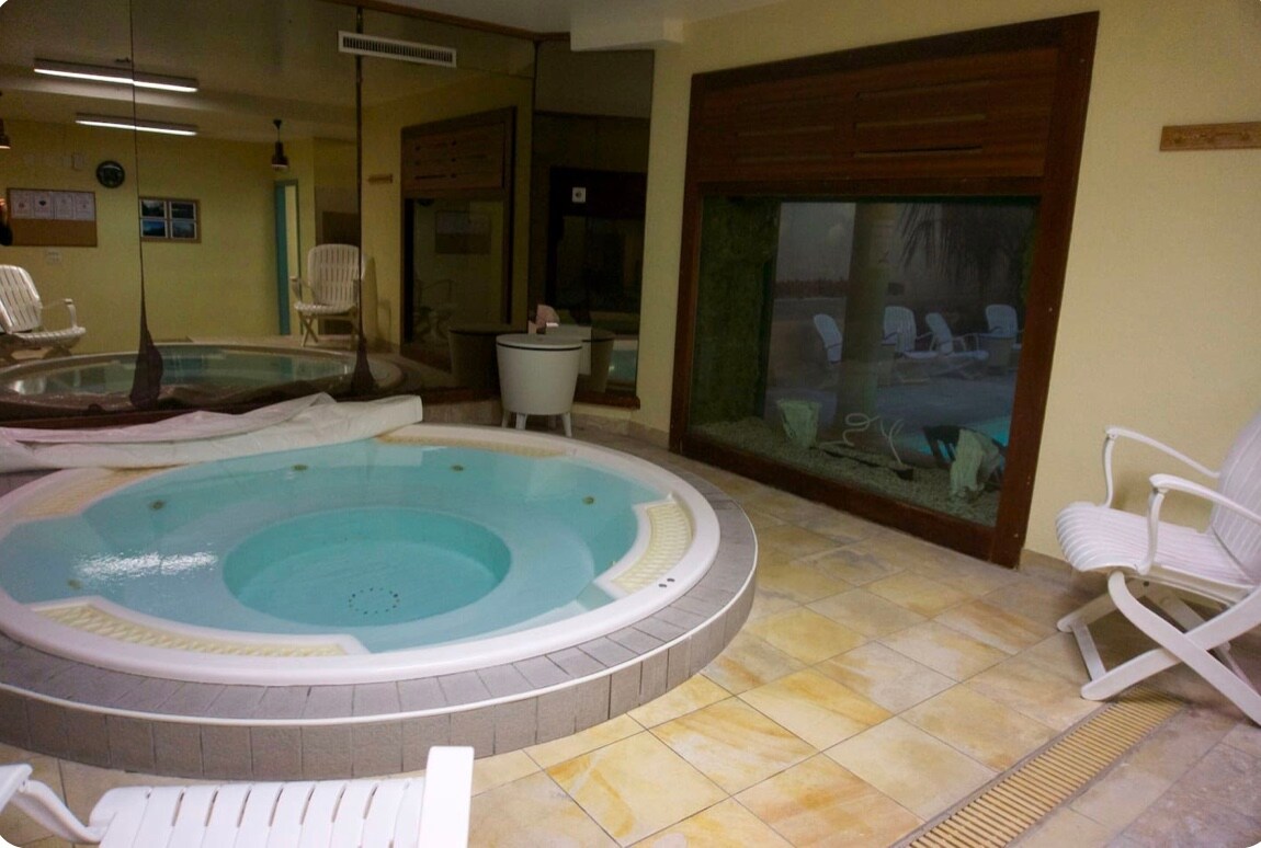 An indoor wellness area features a circular jacuzzi surrounded by stone tile flooring. White lounge chairs are positioned nearby, and a large glass wall reveals a serene view of an aquarium filled with aquatic plants and gentle lighting.