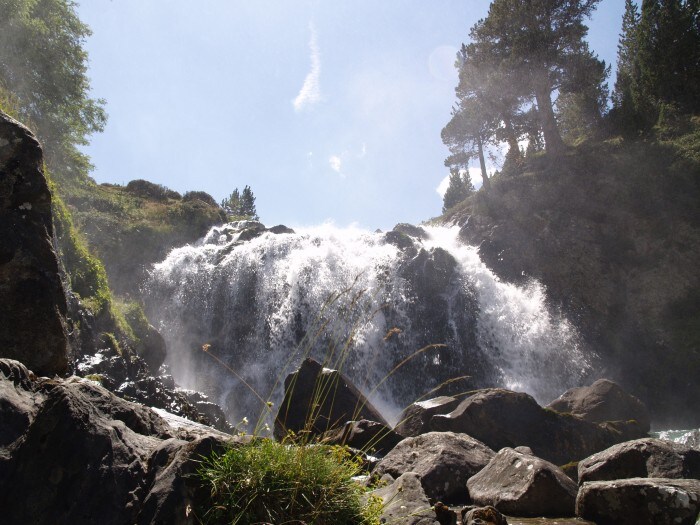 A cascading waterfall is captured amidst a lush, green landscape. Water flows over rocky formations, creating mist in the air. Sunlight filters through the trees above, illuminating the scene and enhancing the vibrant colors of the surrounding nature.