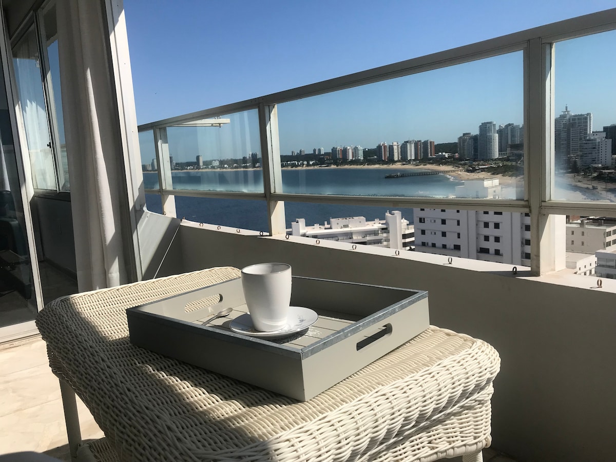 A balcony view showcases a scenic panorama of both Brava and Mansa beaches. A light-colored wicker table holds a tray with a white cup, inviting a moment of relaxation. The clear skies enhance the brightness of the space, contributing to a tranquil atmosphere.