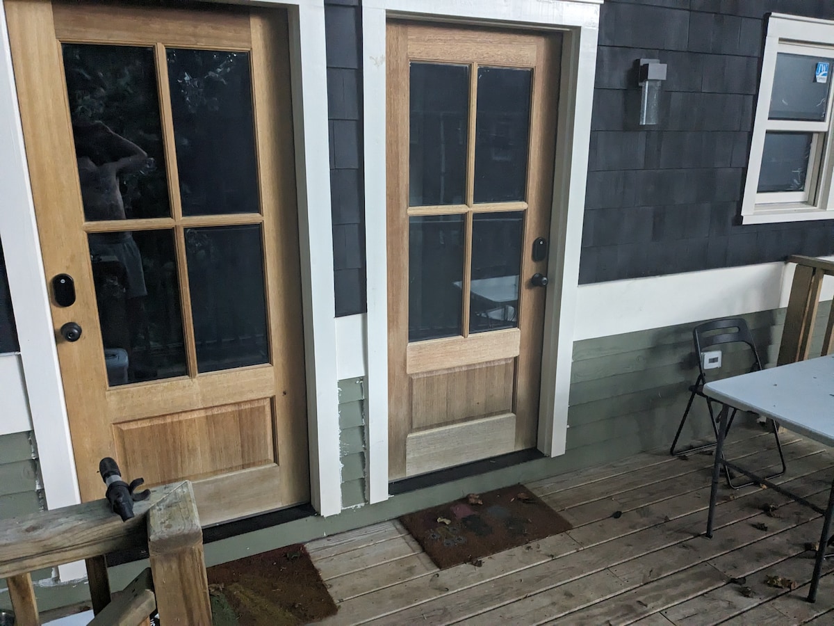 Two wooden doors with glass panels are positioned side by side on a striped wooden deck. The doors feature a minimalist design with four square panes each. A small step leads up to a welcome mat in front of each door, adding a touch of homey charm.