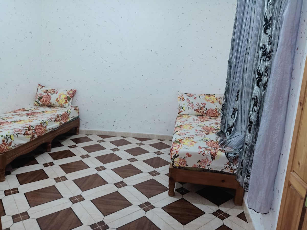 The sleeping area features two twin beds adorned with floral patterned bedding. A light-colored curtain hangs beside the beds, allowing natural light to filter gently into the room. The tiled floor is designed with a geometric pattern, adding visual interest to the space.