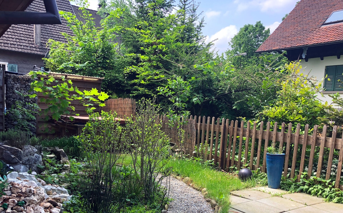 A peaceful private garden is visible, featuring lush greenery and a winding pathway. Various plants and a wooden fence add to the natural ambiance. A stone area and a decorative blue pot can also be seen, inviting relaxation and outdoor time.