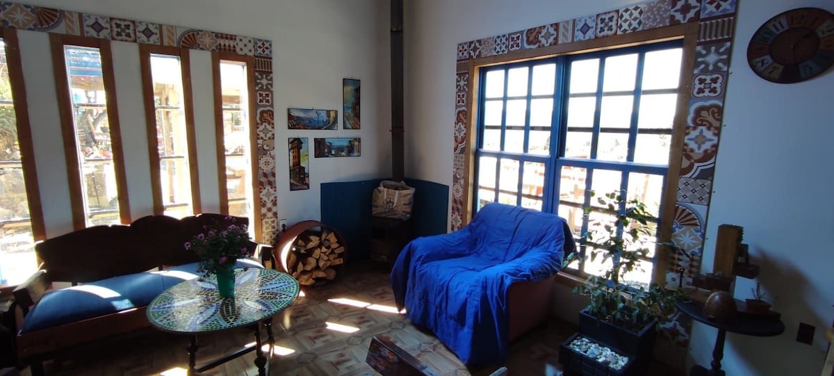 A bright living area showcases large windows framed in blue, allowing natural light to illuminate the space. A comfortable sofa, draped with a blue blanket, sits beside a decorative table adorned with a vase of flowers. Artistic wall accents and a pile of logs add texture.