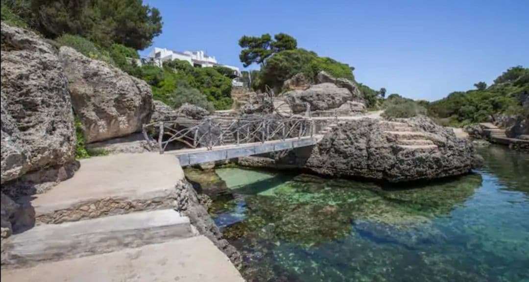 A wooden bridge extends over clear waters, connecting rocky outcrops. The tranquil scene is framed by lush greenery and coastal features, showcasing a serene environment ideal for relaxation. The hillside in the background is dotted with white structures, harmonizing with the natural landscape.