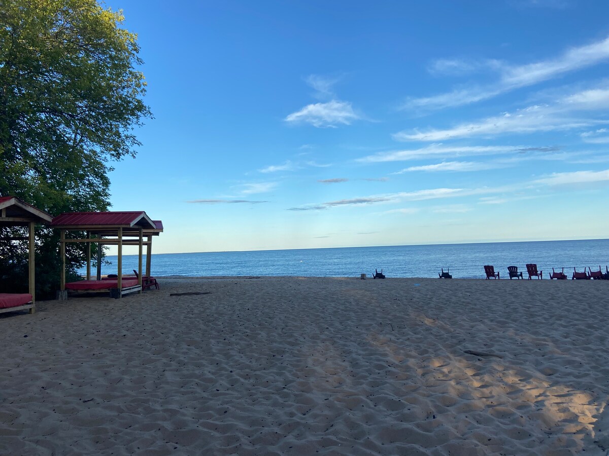 A sandy beach is seen beside a calm lake, with lounge chairs arranged in the foreground. A covered seating area is visible to the left, framed by trees. The sky is clear, showcasing subtle cloud formations.