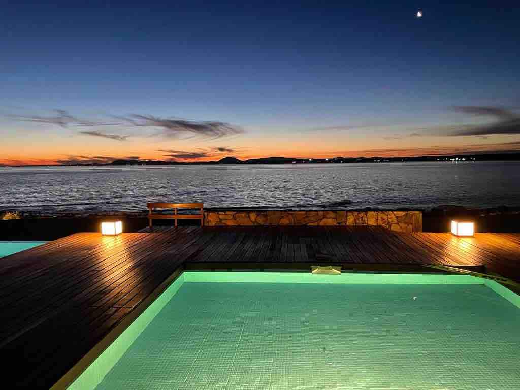 The image captures a serene scene of a pool illuminated in warm tones, overlooking the ocean at dusk. The horizon displays a blend of soft colors as the sun sets, with distant hills and lights reflecting on the water's surface.