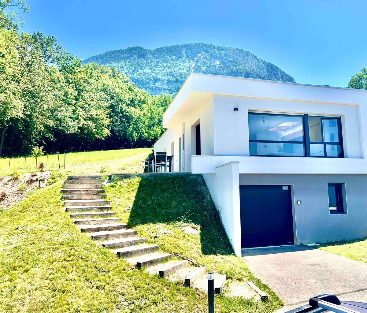 A modern villa is set against a backdrop of lush greenery and mountains. Steps lead from the grassy area to the entrance, where outdoor seating is visible. The structure features large windows and a minimalist design that seamlessly integrates with the natural landscape.
