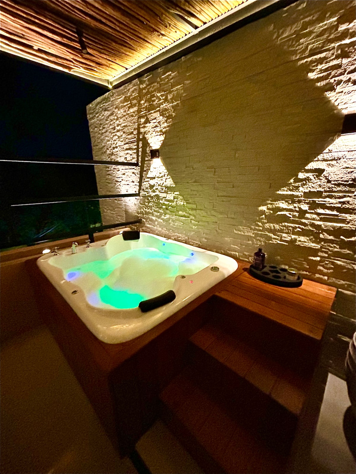 A rooftop jacuzzi is illuminated with soft, multicolored lights, creating a serene ambiance. The spa features a wooden deck leading up to it, with textured stone walls surrounding the area. Subtle lighting enhances the tranquil setting, perfect for relaxation.