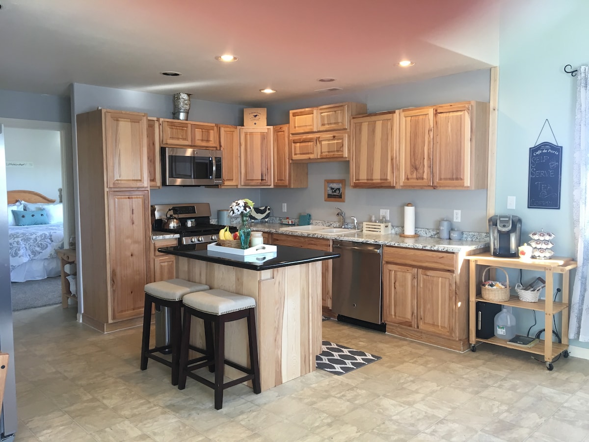 A modern kitchen features natural wood cabinetry and stainless steel appliances. An island with seating is centrally located, and a coffee maker is visible on the counter. Light-colored flooring complements the bright and welcoming atmosphere. A glimpse of the sleeping area is visible in the background.