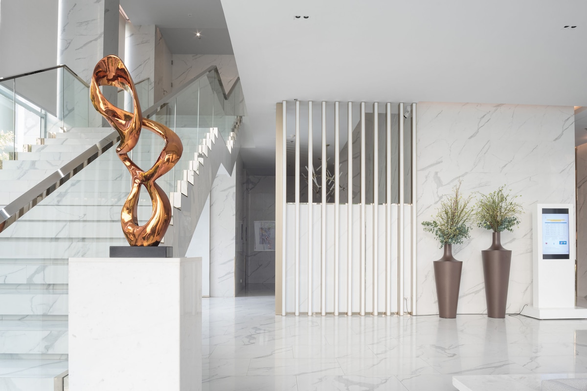 The spacious lobby features elegant marble flooring and modern staircase. A striking bronze sculpture is centered on a white pedestal, while large planters with greenery are positioned on either side. A discreet kiosk is visible against the wall, enhancing the contemporary aesthetic.