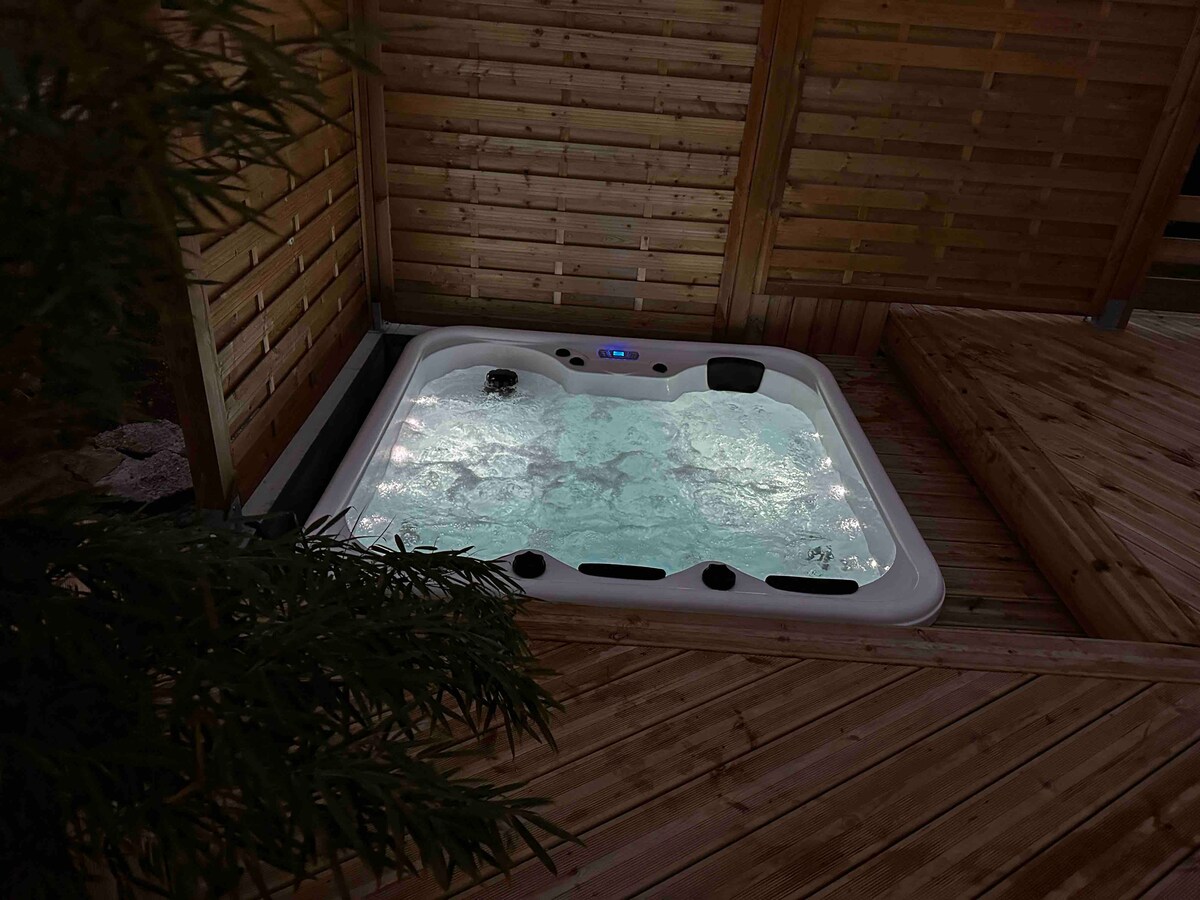 A private jacuzzi for four is situated within a wooden enclosure, surrounded by natural wood paneling. Soft, ambient lighting reflects off the water's surface, enhancing the serene environment. The deck features smooth wooden flooring, providing a comfortable area around the jacuzzi.