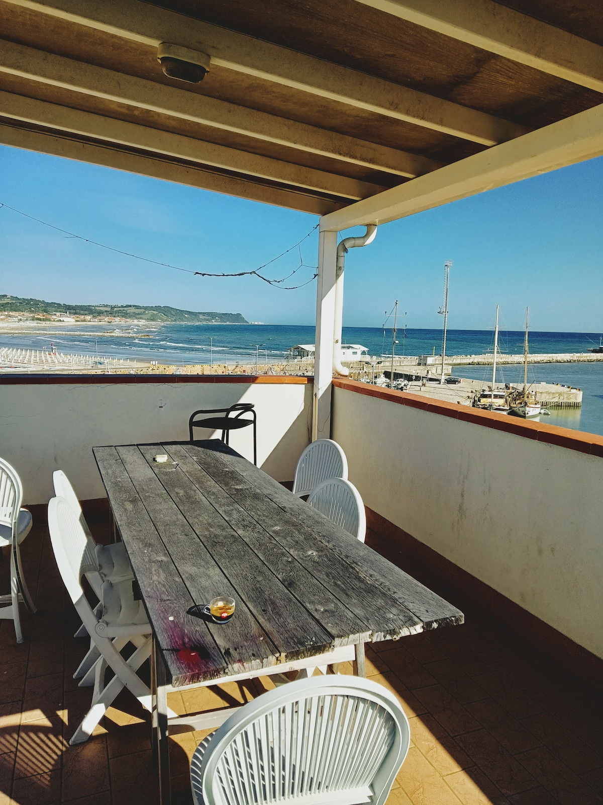 A communal terrace features a large wooden table surrounded by white chairs, offering views of the harbor and the sea. The serene landscape includes boats moored in the water and a coastal backdrop, with sunlight illuminating the outdoor space.