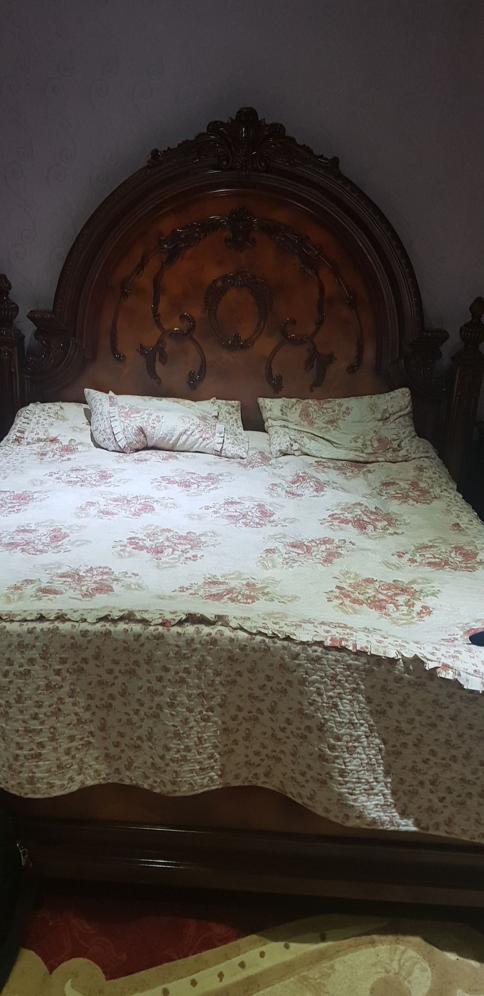 A large wooden bed is adorned with a floral-patterned duvet cover. Multiple decorative pillows are arranged on the bed. The elaborate headboard features intricate detailing, contributing to a traditional aesthetic in the room.