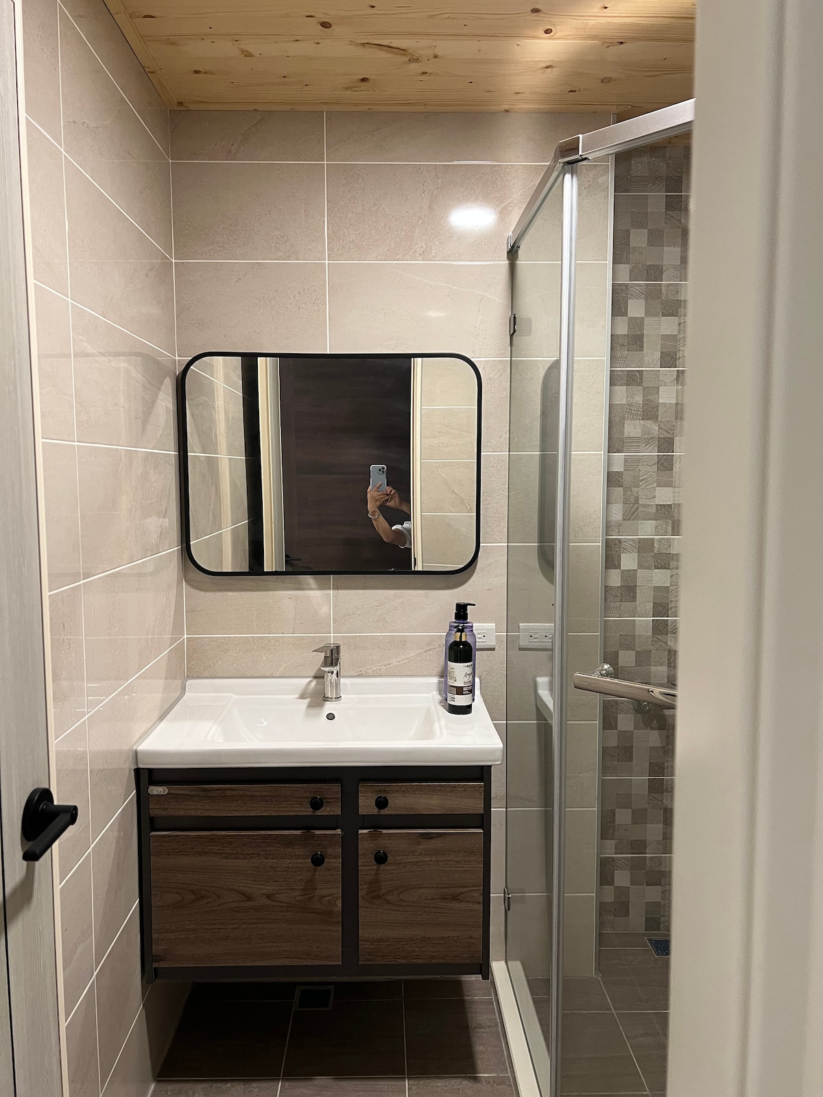 A modern bathroom showcases a wall-mounted mirror above a sleek vanity with a sink and wooden storage drawers. A glass shower stall with patterned tiles is visible to the right, and neutral-toned tiles adorn the walls, contributing to a clean and spacious appearance.