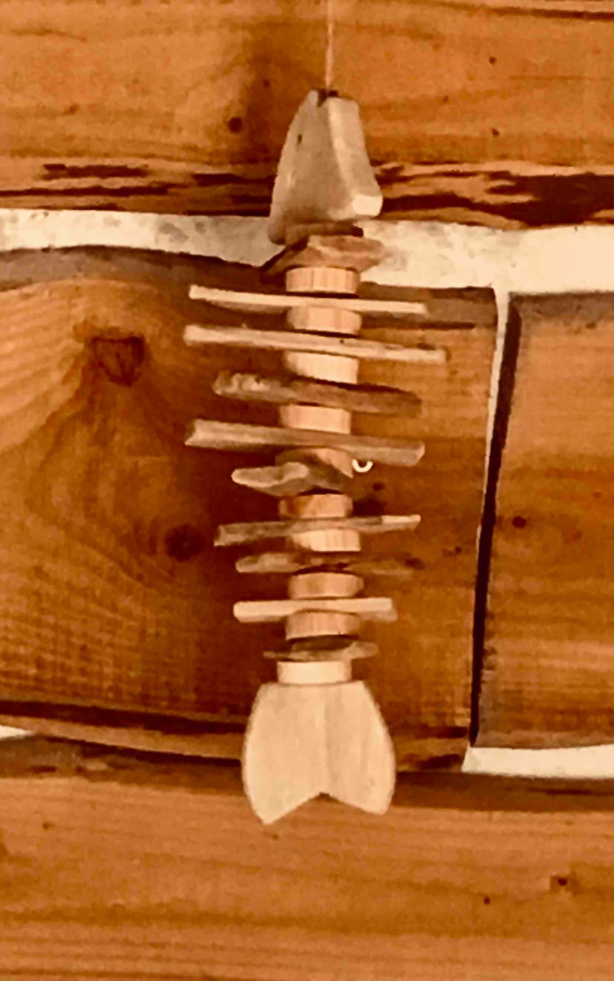 A wooden fish sculpture is displayed against a rustic wooden wall. The sculpture features a series of stacked wooden pieces forming the shape of a fish, highlighting the craftsmanship in its design.