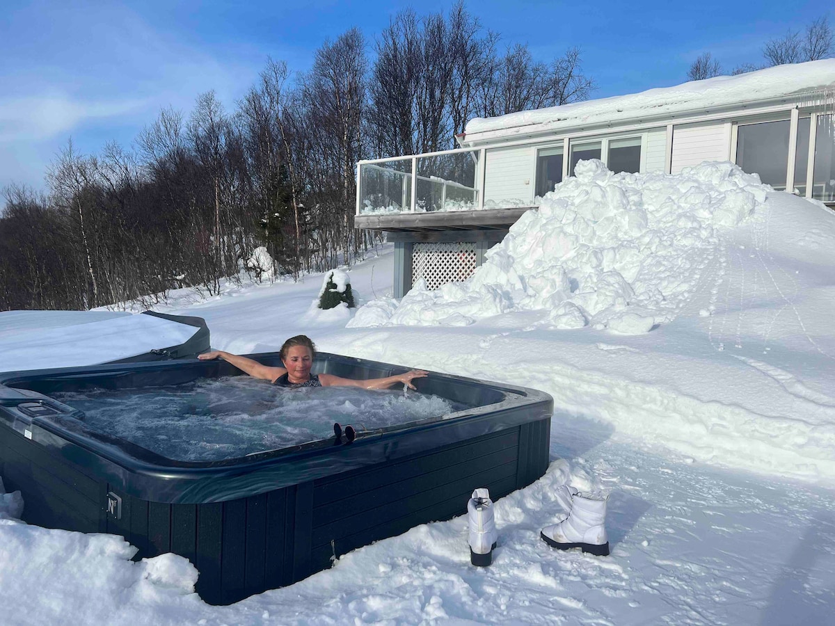 A jacuzzi is seen surrounded by a snowy landscape, with a house in the background. The clear water of the jacuzzi contrasts with the white snow, while footprints lead towards the hot tub. Snowdrifts rise around the area, creating a serene winter setting.