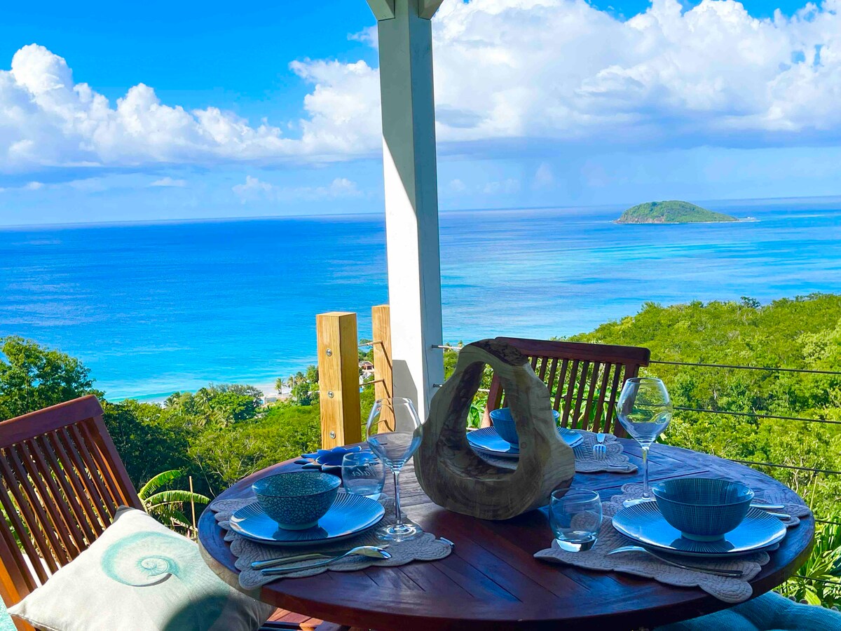 A dining table is set for a meal, featuring blue dishware and glassware. The table is positioned on a terrace overlooking the ocean, with scenic views of the water and greenery. Lush hills are visible beyond, framed by a clear blue sky and scattered clouds.