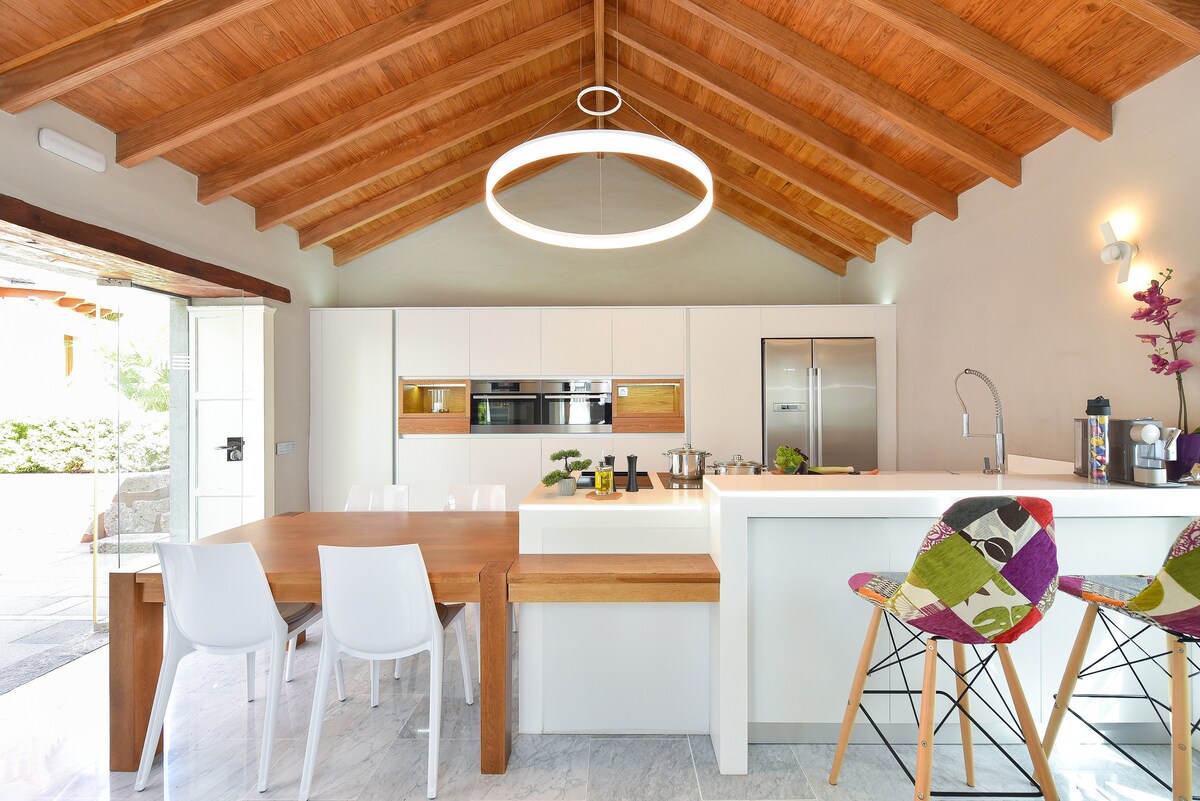 The kitchen area features sleek, modern cabinetry and stainless-steel appliances. A central island with seating is complemented by a wooden dining table. High wooden beams create a spacious atmosphere, while a circular light fixture adds contemporary illumination.