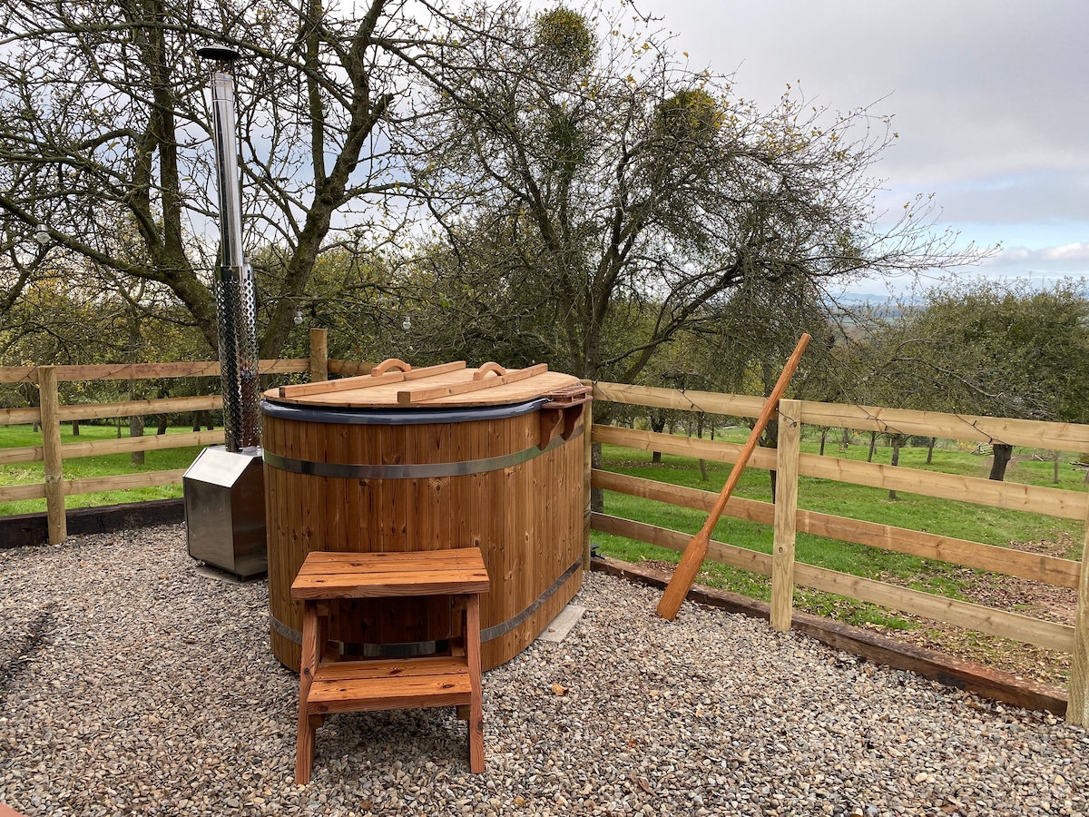 A wood-fired hot tub is situated outdoors, surrounded by a wooden fence and gravel flooring. A small wooden bench is positioned nearby. Trees and a green landscape are visible in the background, contributing to a serene outdoor setting.