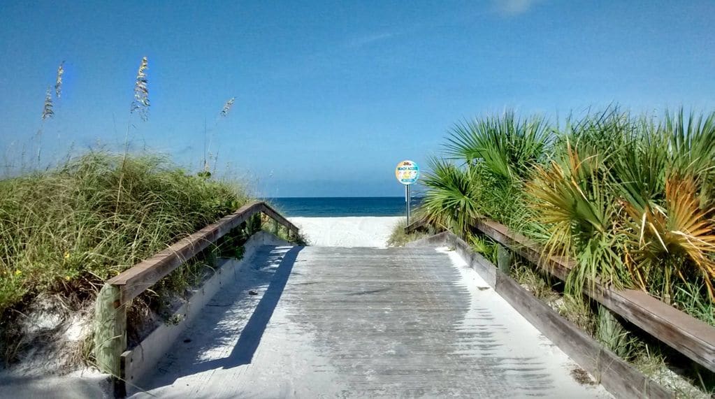 A wooden boardwalk leads directly to the beach, surrounded by tall grasses and lush palm trees. The serene ocean view features calm waters under a clear blue sky, with a beach access sign visible in the distance. The sandy pathway is inviting and well-maintained.