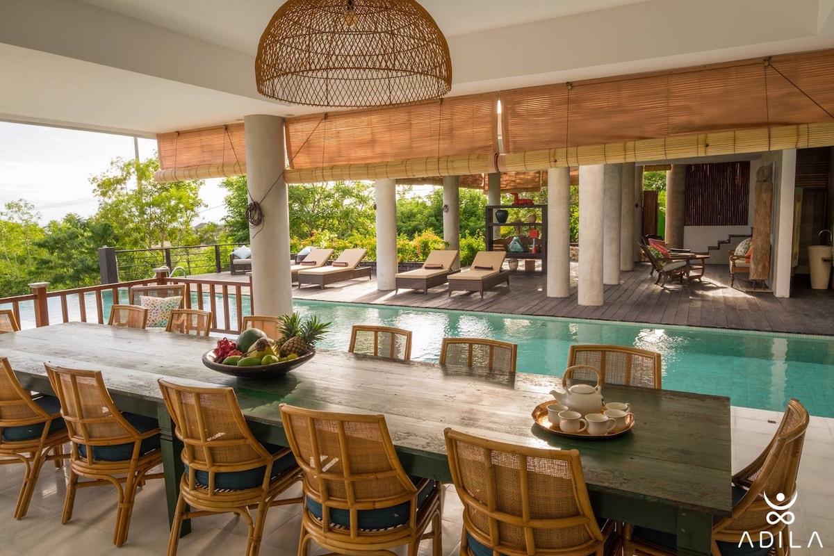 An open dining area features a long, rustic table surrounded by ten rattan chairs. Large windows overlook a pool and lush greenery, while natural light brightens the space. The setting is enhanced by a high, woven ceiling, creating a spacious and inviting atmosphere.
