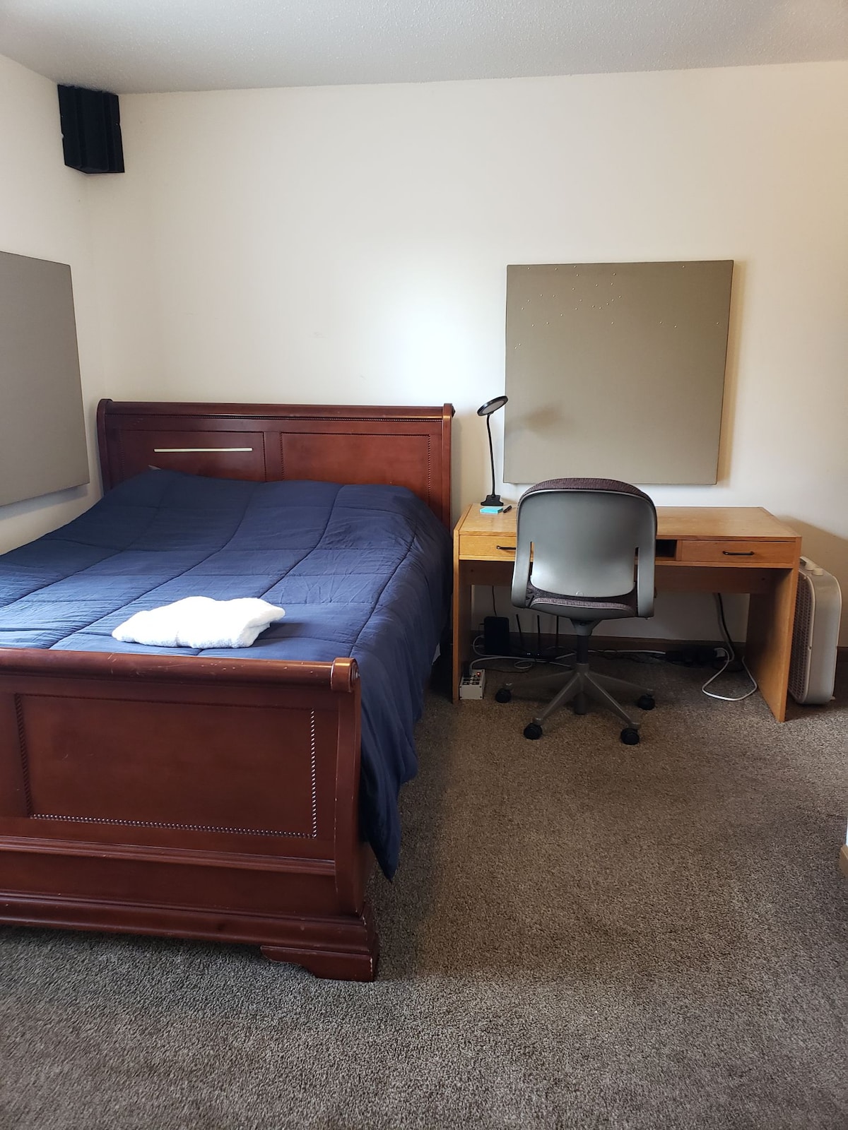 A small bedroom features a wooden bed covered with a navy blue blanket, complemented by a white towel. A desk with a wheeled chair is placed next to the bed, and a desk lamp is positioned on the surface. Two muted wall panels provide a neutral backdrop.