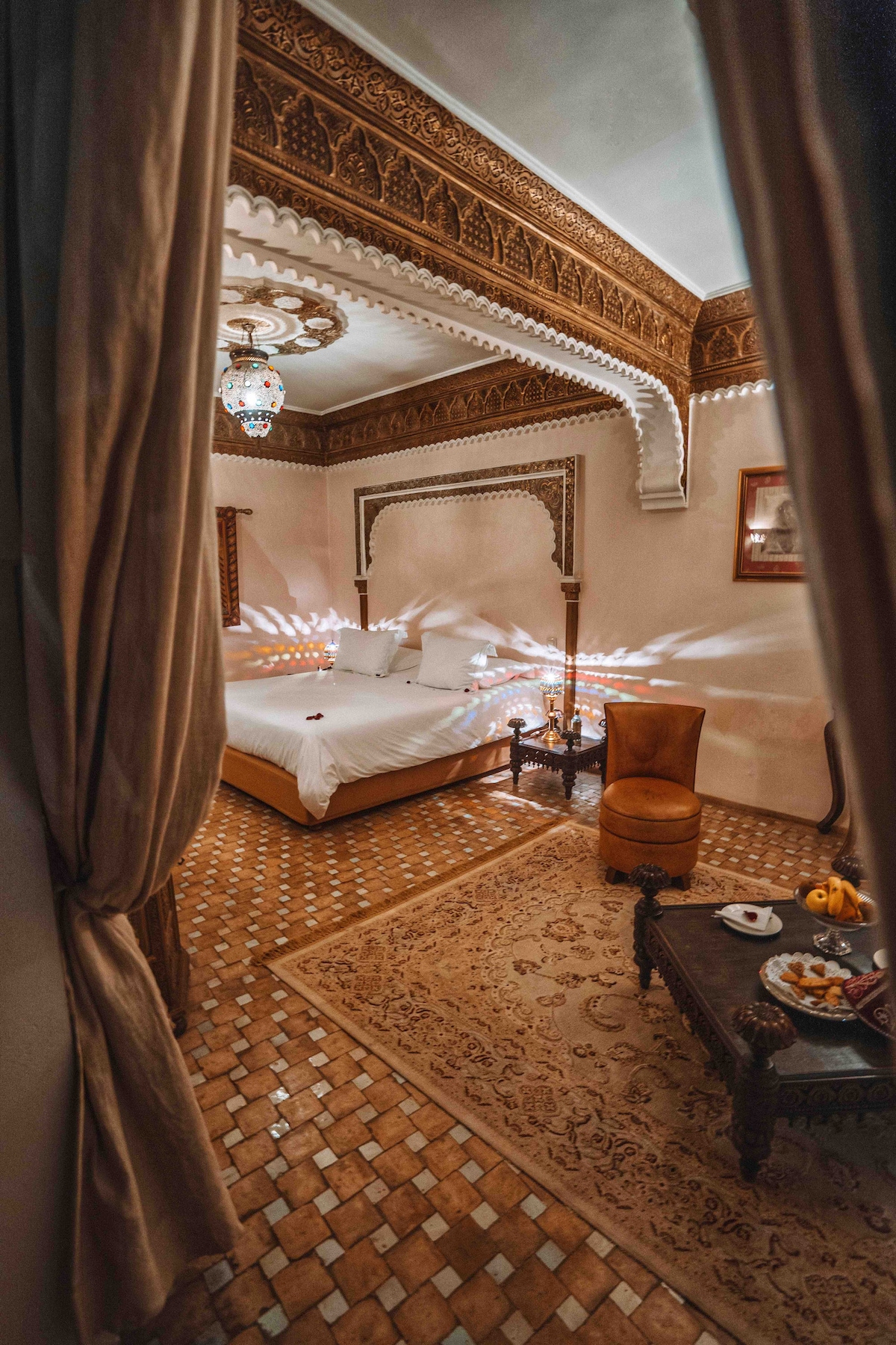 A spacious room showcases traditional Moroccan architecture with intricate woodwork and elegant lanterns. A comfortable bed is centrally placed, accompanied by a small, decorative table and a cozy armchair. Natural light filters through, creating a serene atmosphere, with a patterned rug adorning the tiled floor.