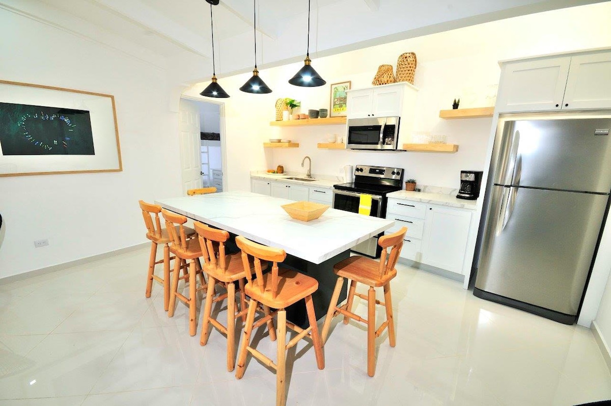 A modern kitchen showcases sleek white cabinetry and stainless steel appliances. A spacious island is surrounded by wooden stools, inviting social gatherings. Natural light enhances the open design, while shelves are adorned with decorative items and kitchen essentials.
