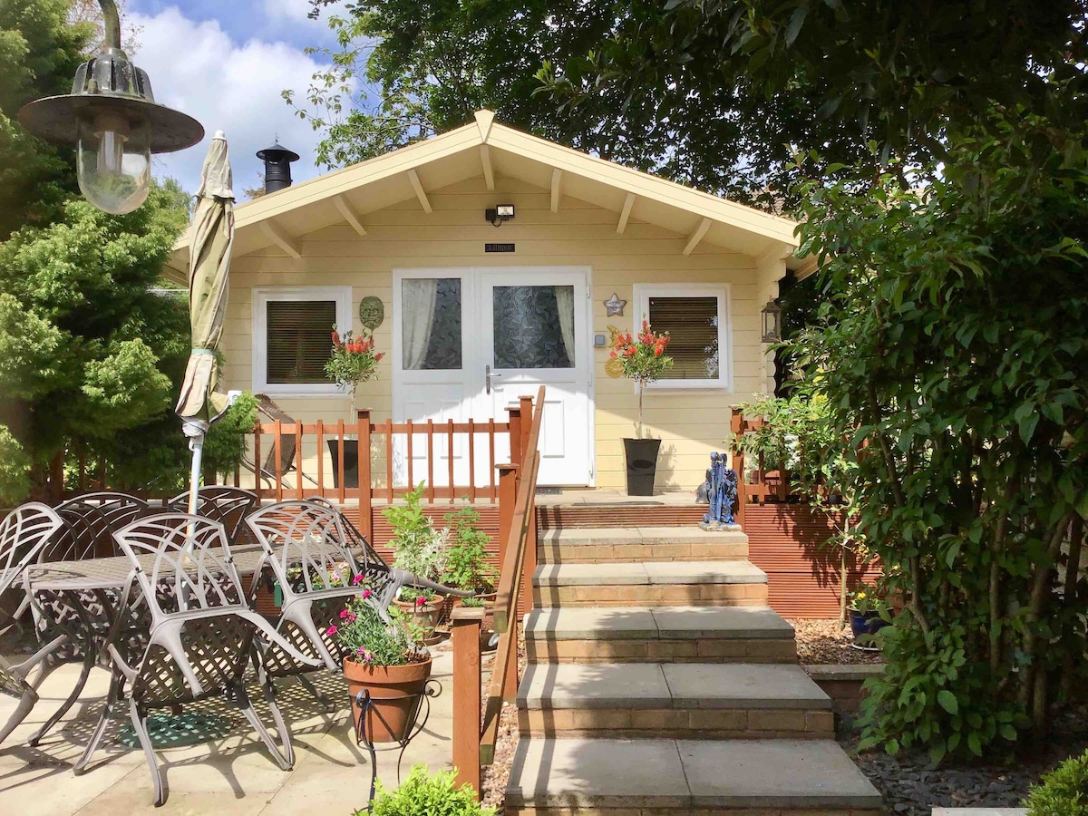The exterior of Lilliput features a wooden log cabin with a welcoming entrance. Steps lead up to a porch adorned with flowers. A patio area accommodates a dining set with several chairs, surrounded by greenery and decorative plants, creating a cozy outdoor space.