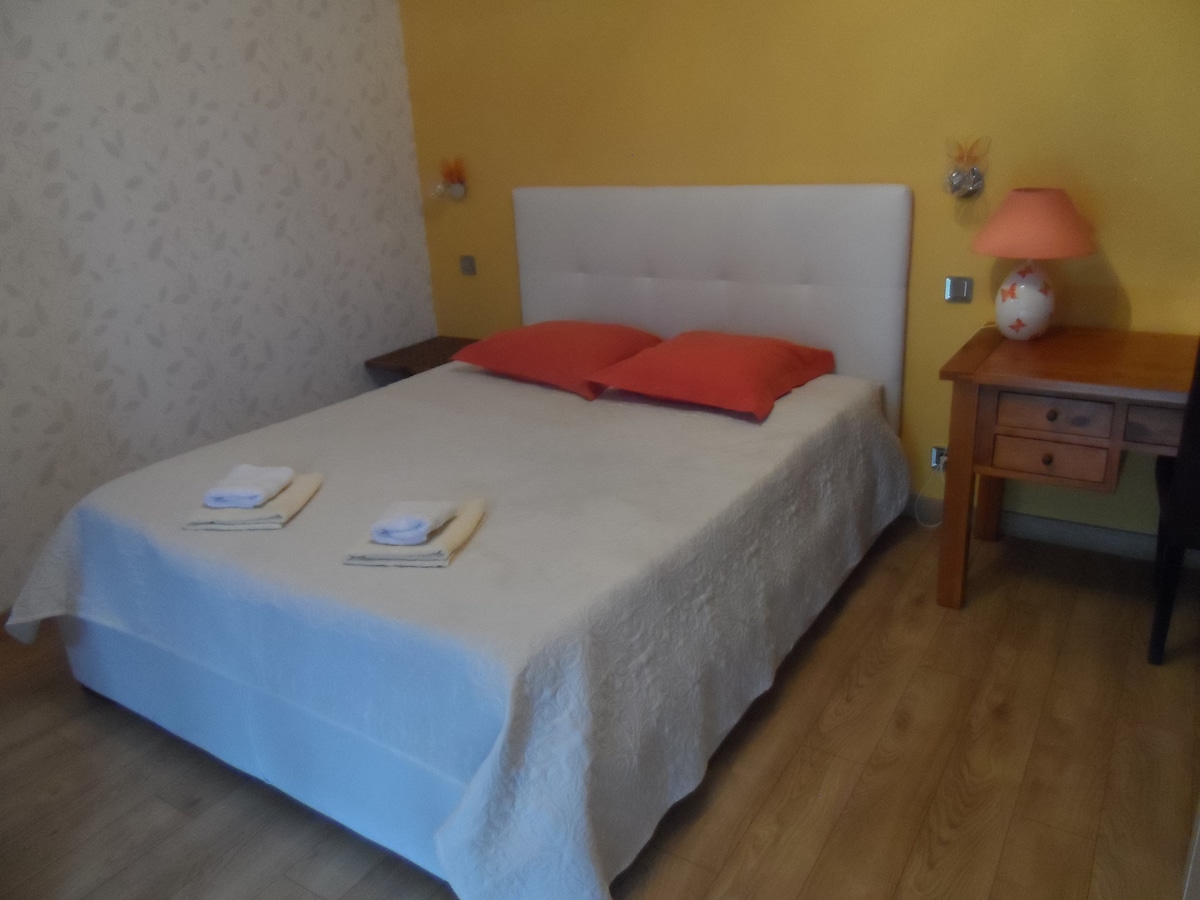 The bedroom features a spacious bed dressed in a light-colored coverlet, complemented by two vibrant orange pillows. A bedside table with a lamp sits beside the bed, while soft wood flooring adds warmth to the space. The walls are adorned with subtle floral patterns against a yellow backdrop.