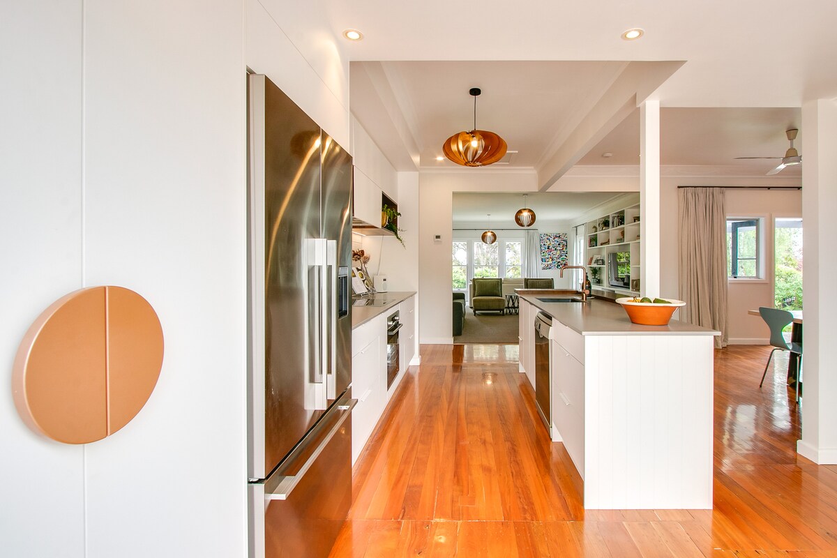 A modern kitchen with a central island and stainless steel appliances is visible. Light-colored cabinetry and warm wooden flooring enhance the spaciousness. Bright pendant lights hang above, and an open plan layout leads towards a comfortable living area in the background.