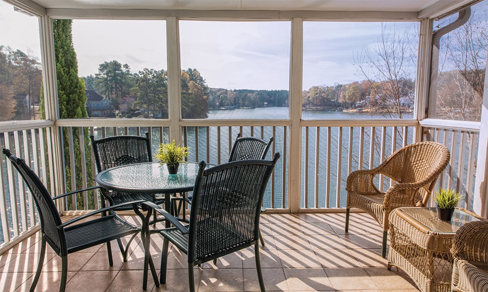 A screened-in porch features a round dining table with four black chairs and a cozy rattan chair. A small potted plant is placed on the table. Sunlight casts shadows on the tiled floor, while a scenic lake view is framed by the porch's railing.